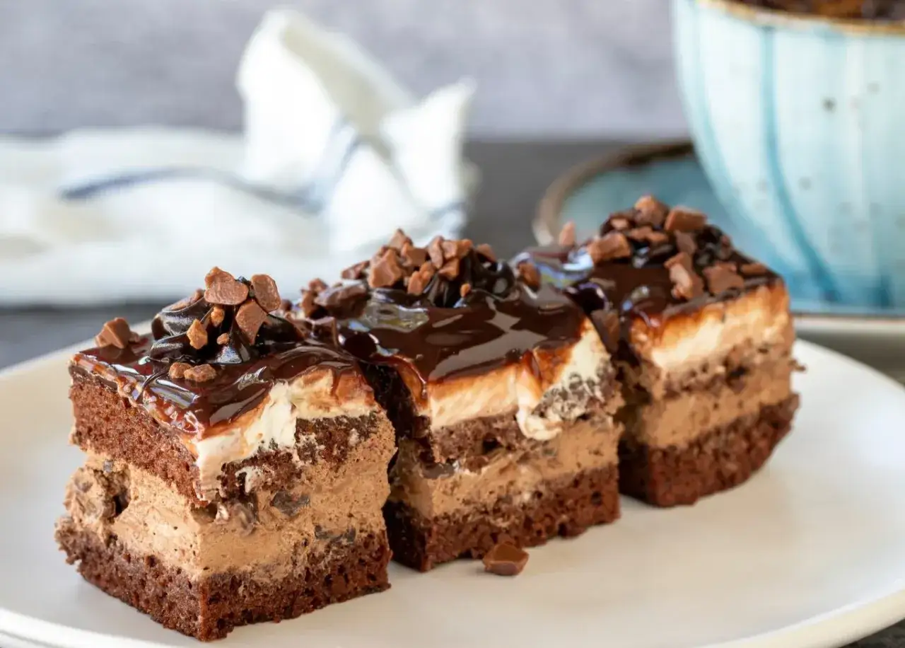Trzy kawałki ciasta Kinder Bueno z mascarpone, polane czekoladą i posypane kawałkami czekolady.