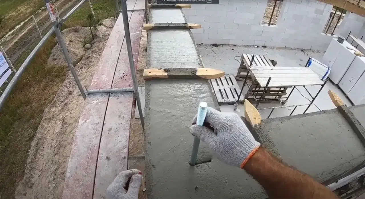 Ręka w rękawicy osadza kotwę w świeżym betonie, przygotowując fundamenty.