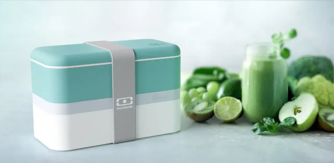 A teal and white bento box sits next to a green smoothie and fresh produce, hinting at how to keep bento box cold for a refreshing meal.