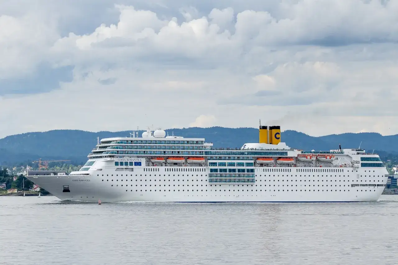 La nave da crociera Costa Romantica naviga in acque calme, con montagne verdi sullo sfondo e un cielo nuvoloso.