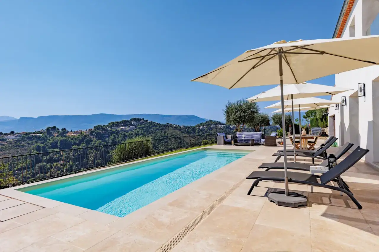 Piscine à débordement sur un terrain en pente, offrant une vue panoramique sur les collines verdoyantes et le ciel bleu. Des chaises longues et des parasols invitent à la détente.