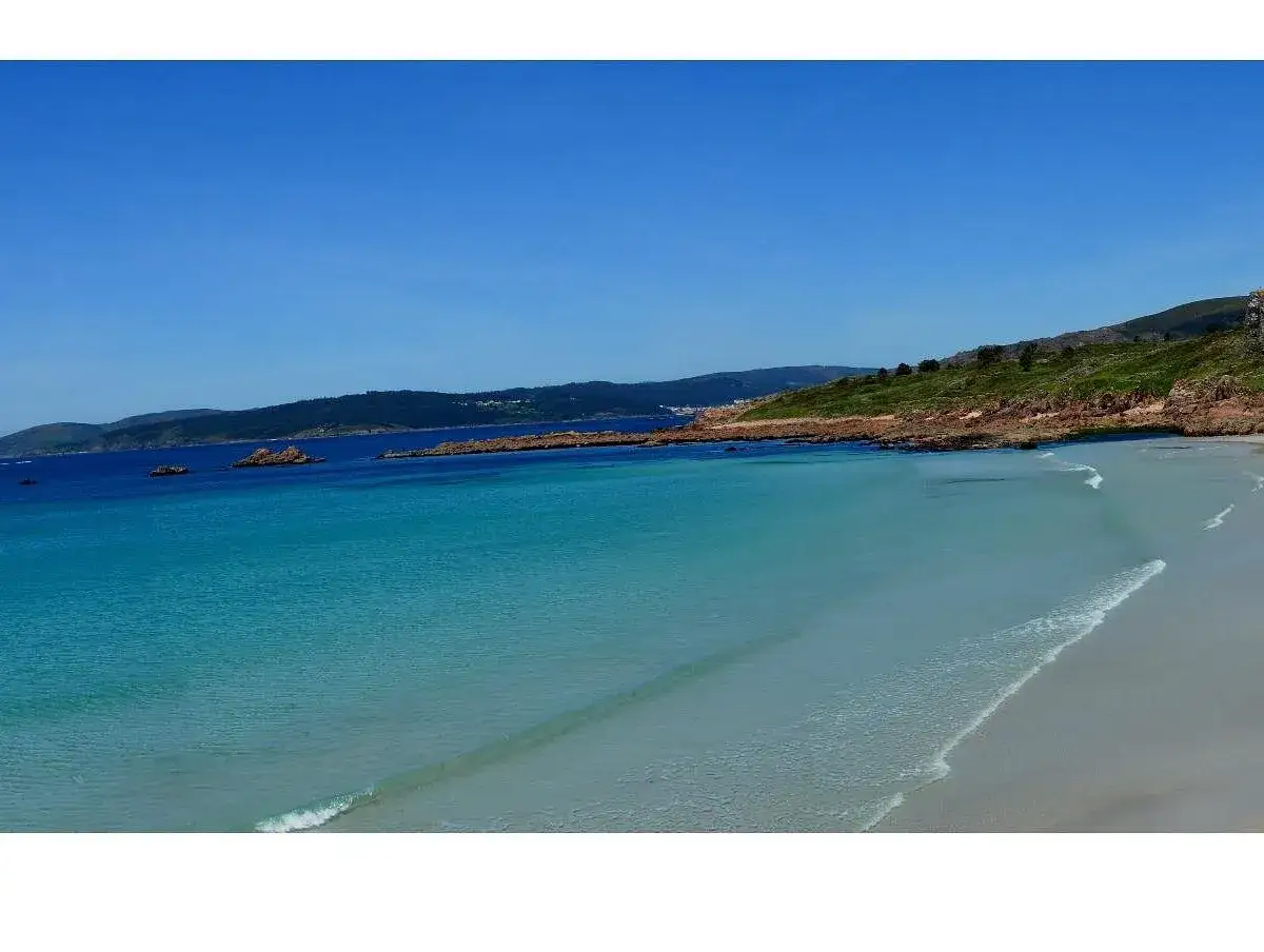 Aguas turquesas y arena dorada en la playa de San Pedro, Galicia. Un paisaje sereno bajo un cielo azul intenso.