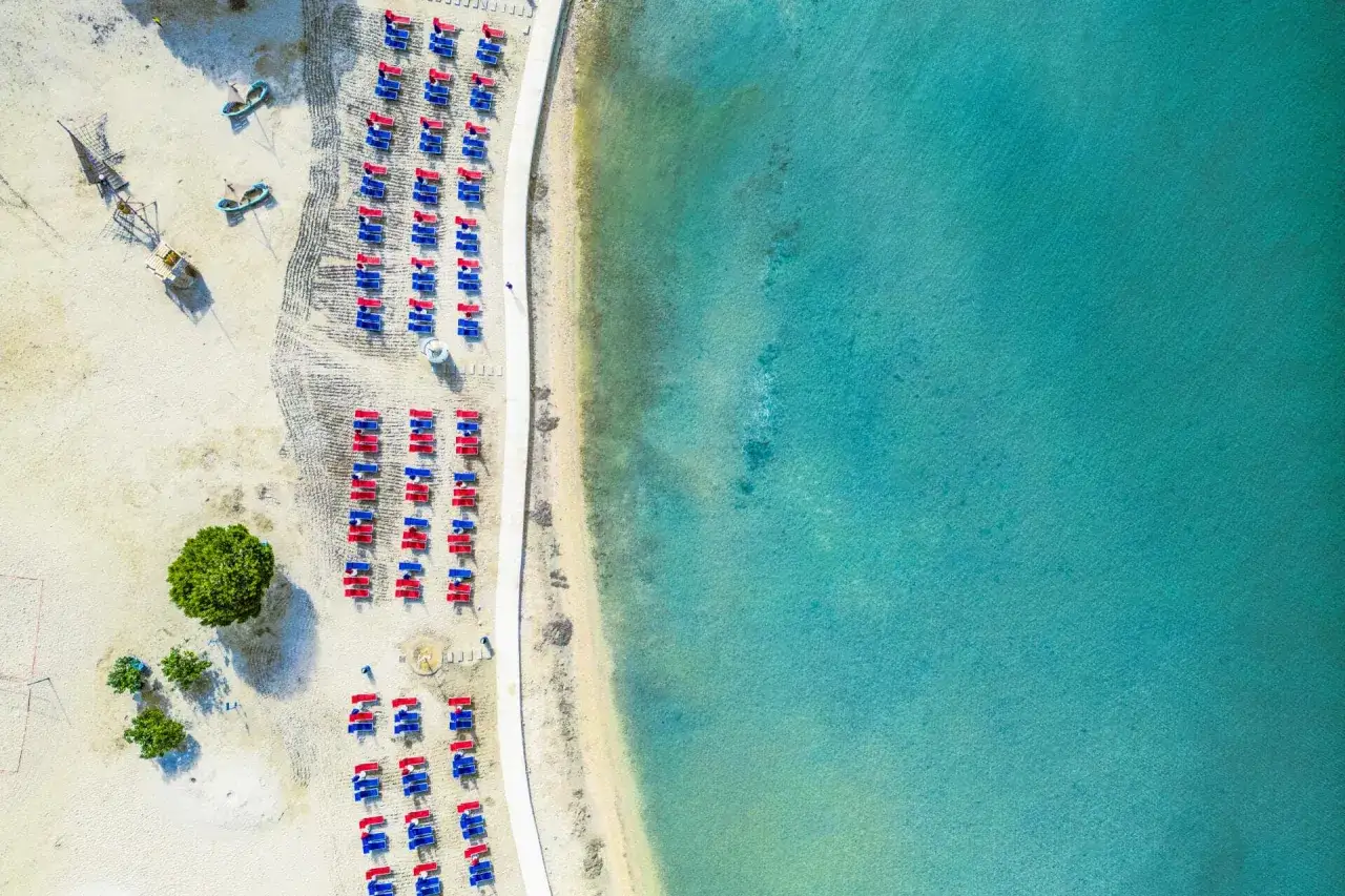 Widok z lotu ptaka na plażę kempingu Zaton: rzędy czerwonych i niebieskich leżaków, czysta woda i piasek.