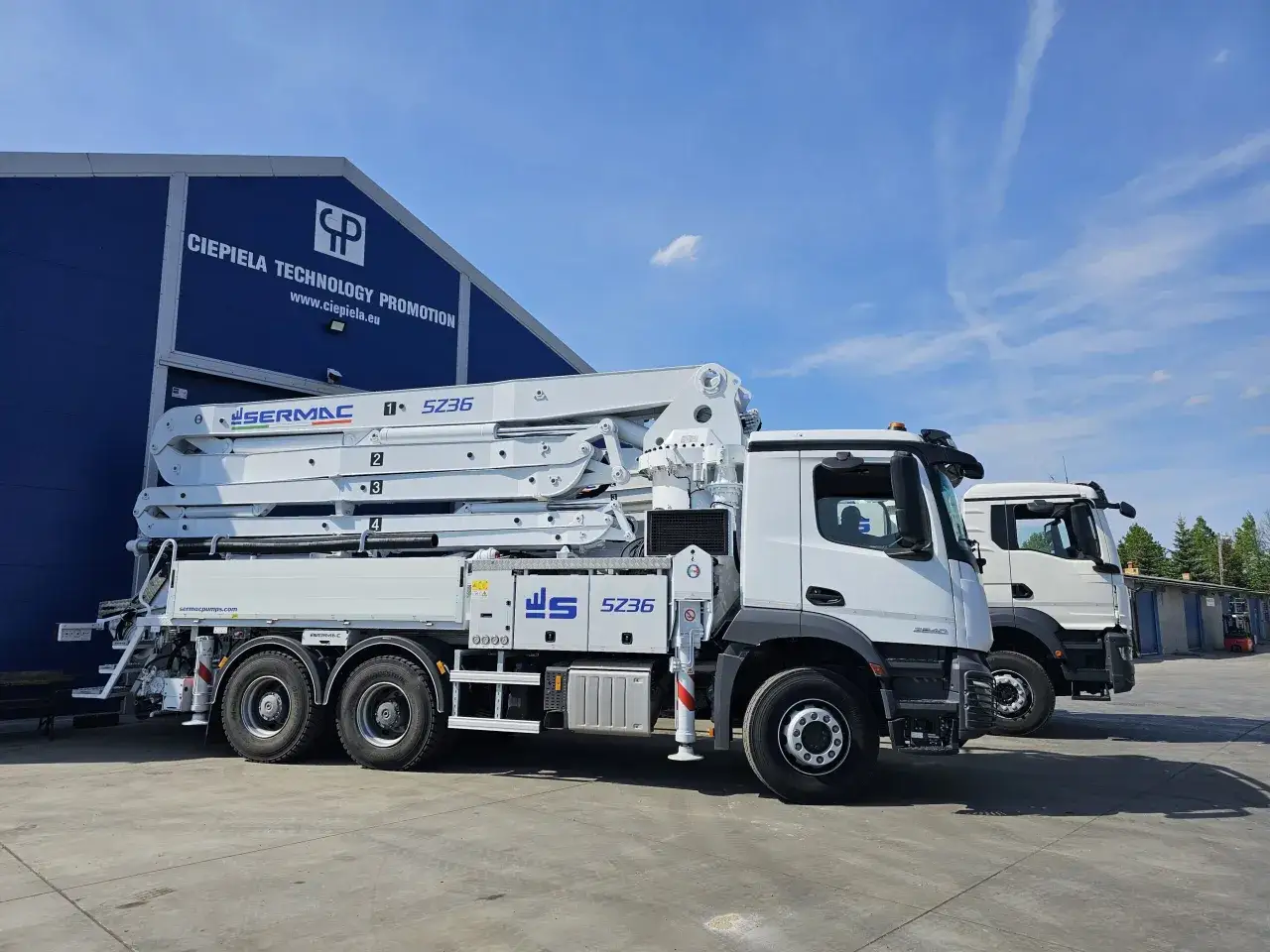 Dwie pompy do betonu Sermac SZ36 stoją przed budynkiem z logo firmy. Zapytaj o to, ile kosztuje kubik betonu, a dowiesz się, jak wiele można przetransportować.