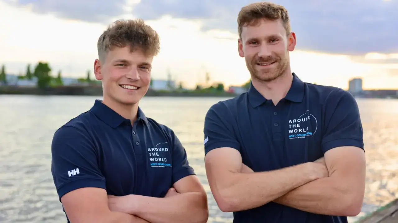 Melwin Fink und sein Teamkollege posieren l&auml;chelnd am Wasser. Sie tragen marineblaue Poloshirts mit dem Aufdruck 