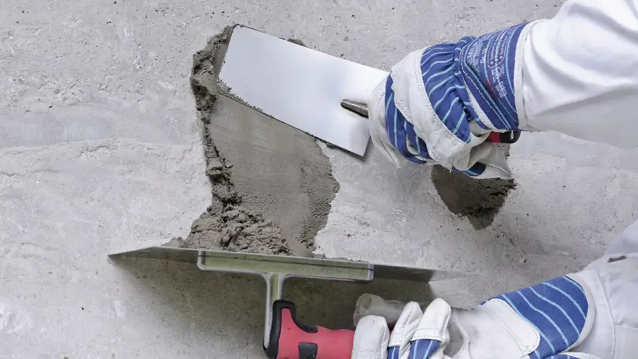 Ręce w rękawicach nakładają masę naprawczą na betonową ścianę za pomocą kielni.