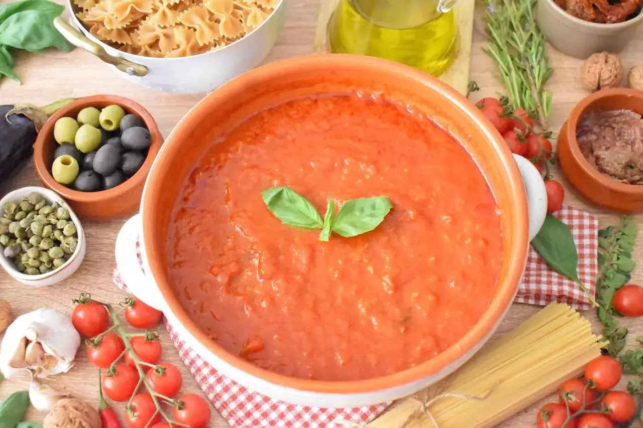 Pyszny, domowy sos do spaghetti w glinianej misie, udekorowany bazylią. Obok składniki: makaron, oliwki, kapary, pomidory, czosnek i oliwa.