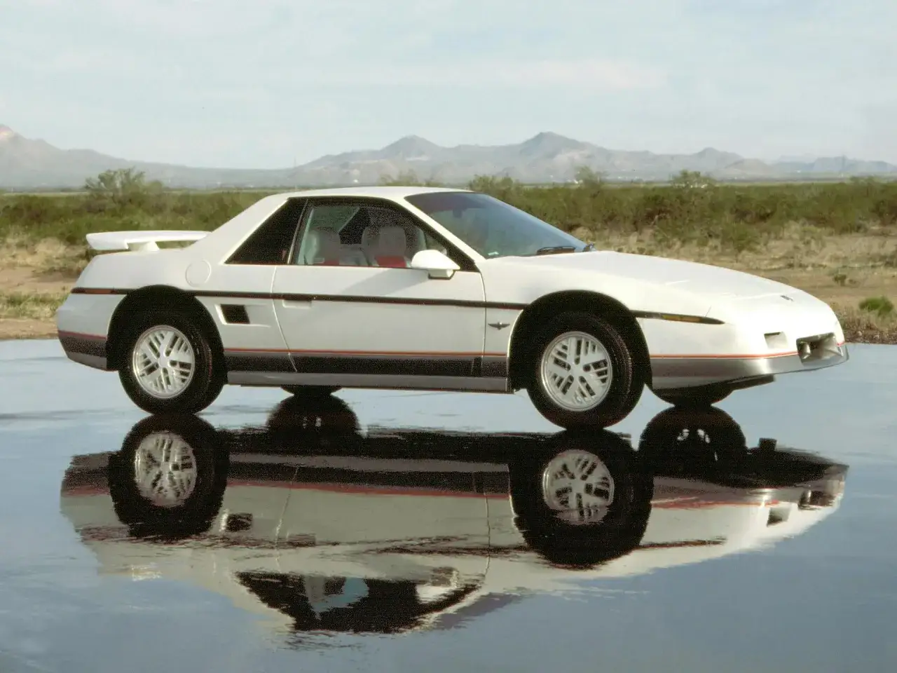 Vit Pontiac Fiero från 80-talet reflekteras i en blank yta, med berg i bakgrunden.