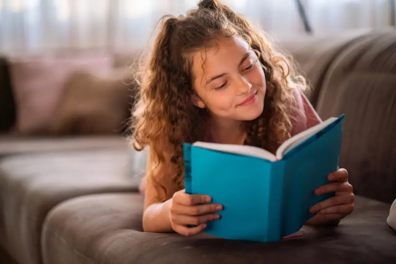 Ein Mädchen liegt auf einem Sofa und liest ein blaues Buch. Sie strahlt und zeigt, wie sie eigenverantwortliches Lernen genießt.