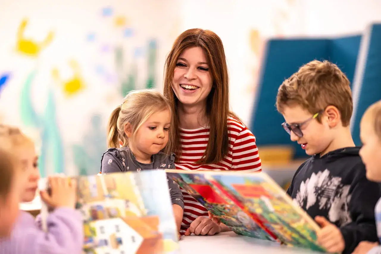 Eine junge Frau mit roten Haaren liest Kindern vor. Sie l&auml;chelt fr&ouml;hlich. Die Kinder sind aufmerksam. Vielleicht denkt sie &uuml;ber ihre PIA Ausbildung und das Gehalt netto nach.