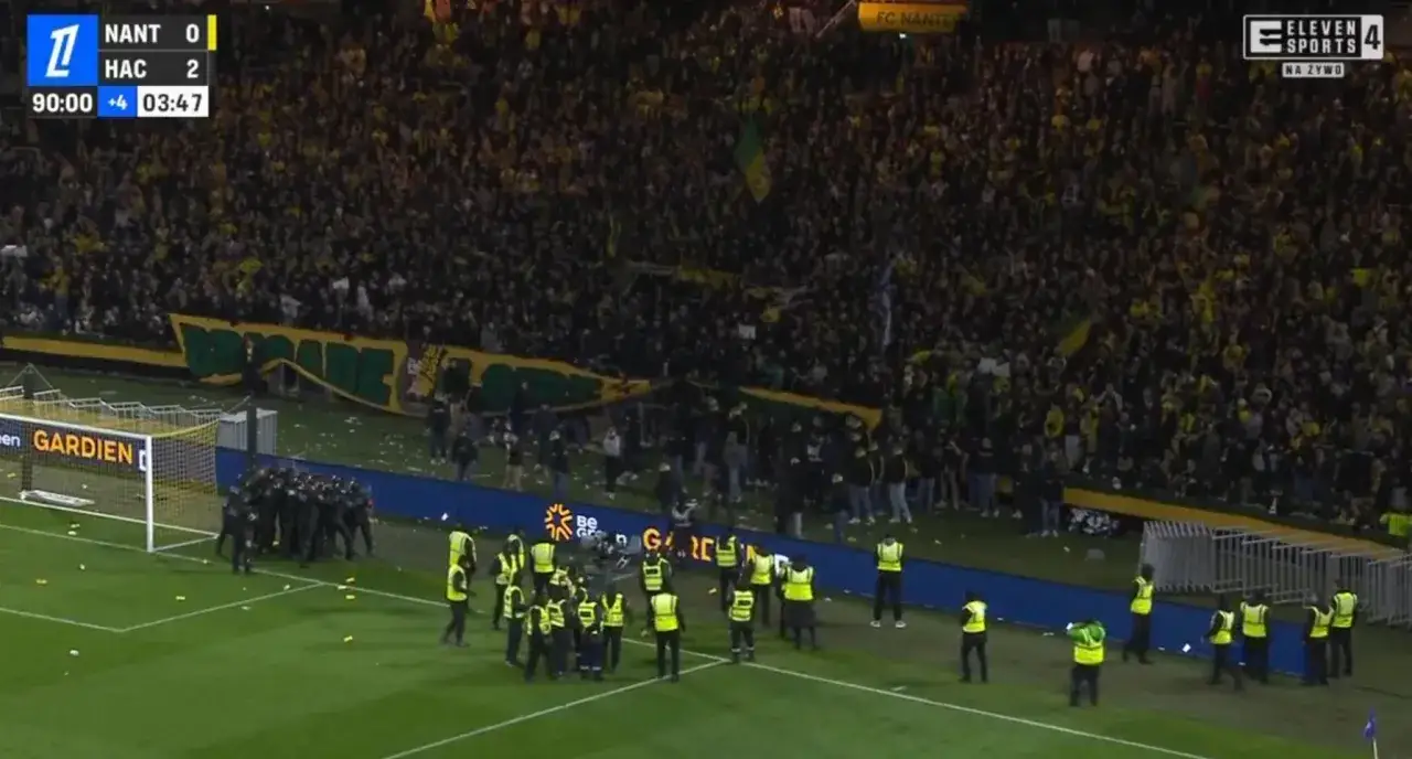 Tłum kibiców na meczu Ligue 1. Na boisku ochrona otacza grupę osób. Wynik 0:2 dla HAC.
