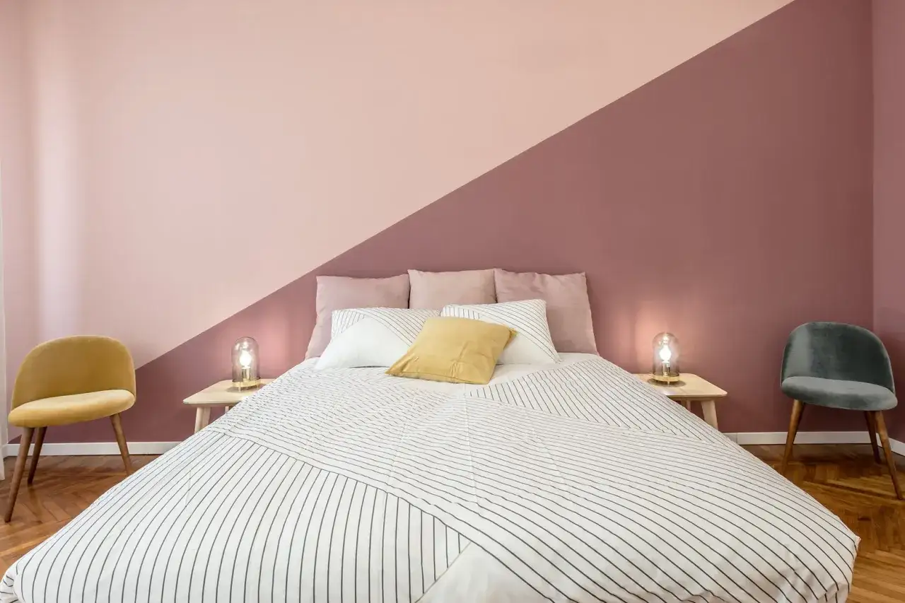 Colori stanza: letto con copriletto a righe, cuscini rosa e gialli, pareti rosa e malva, sedie gialla e grigia.