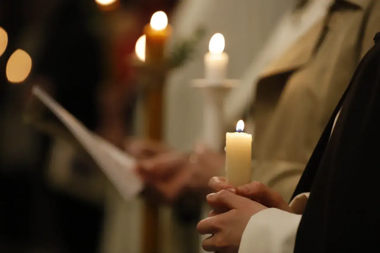 Dłonie trzymają zapalone świece podczas Wigilii Paschalnej. W tle widać innych uczestników i czytania.