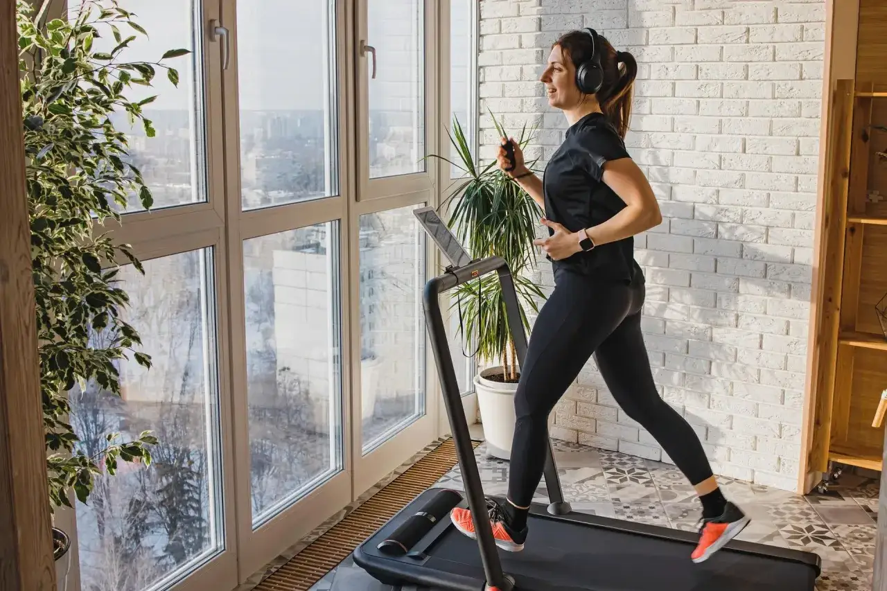 Kobieta w słuchawkach biega na bieżni, zastanawiając się, czy robic cardio na masie. Trening w domu z widokiem na miasto.