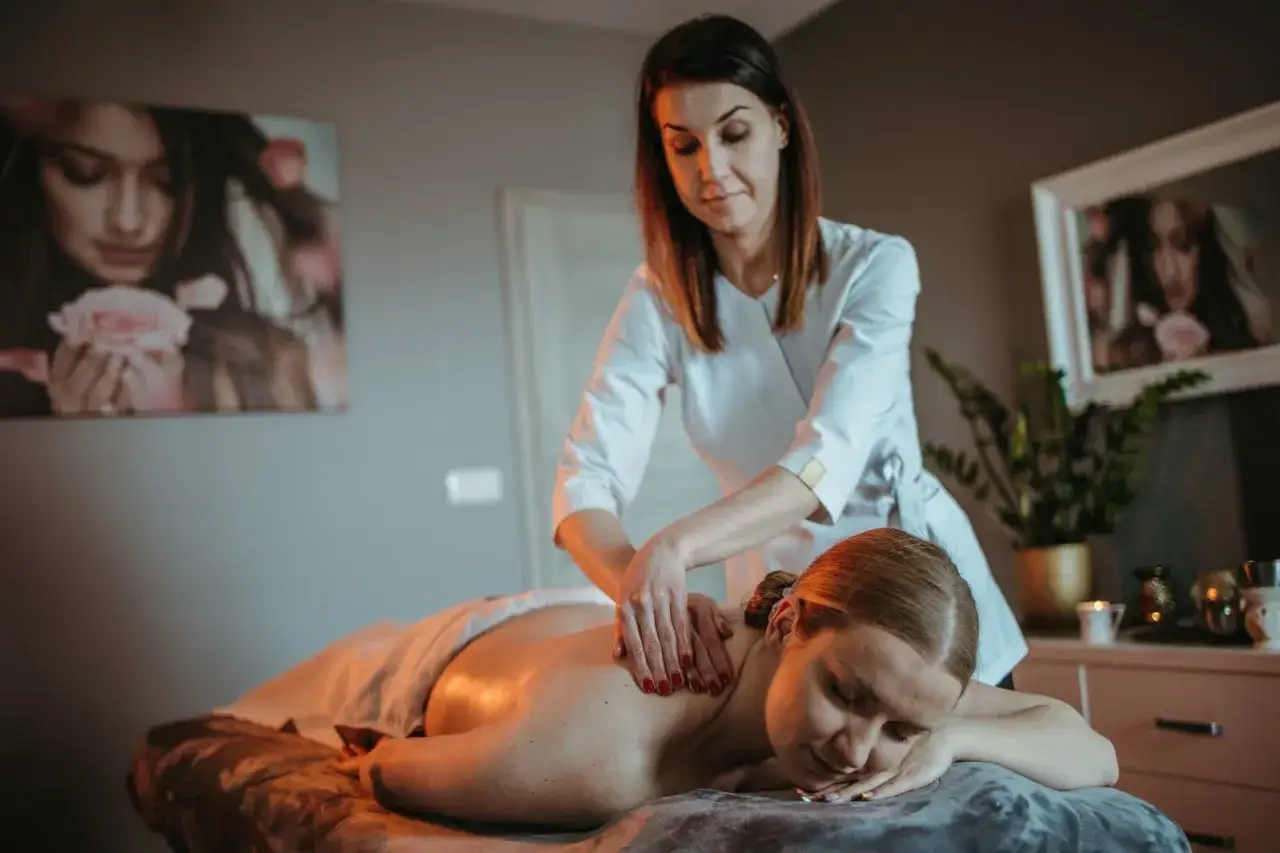 Kobieta otrzymuje **masaż relaksacyjny** pleców. Terapeuta w białej bluzie delikatnie ugniata jej ramiona.