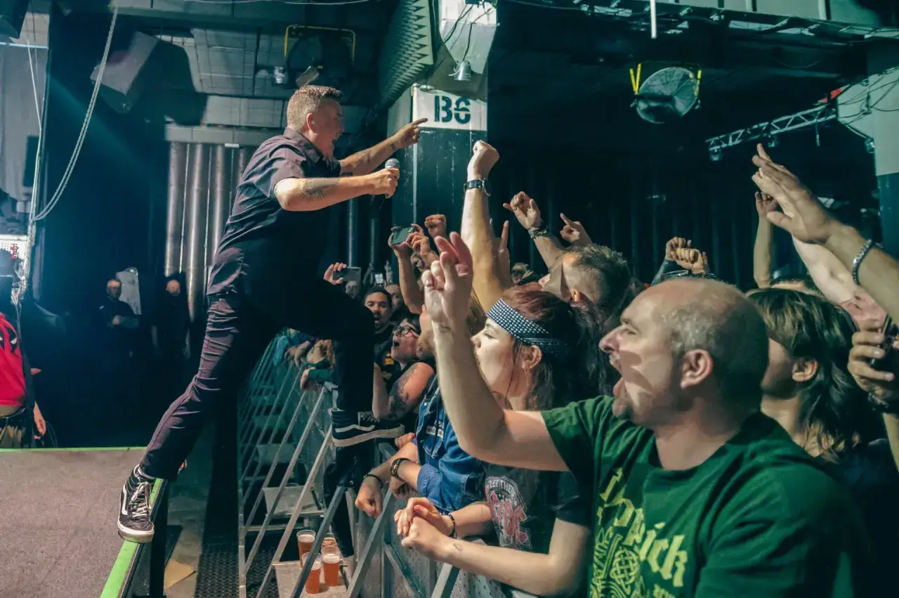 Energiczny wokalista Dropkick Murphys skacze na barierkę podczas koncertu, fani szaleją z radości.