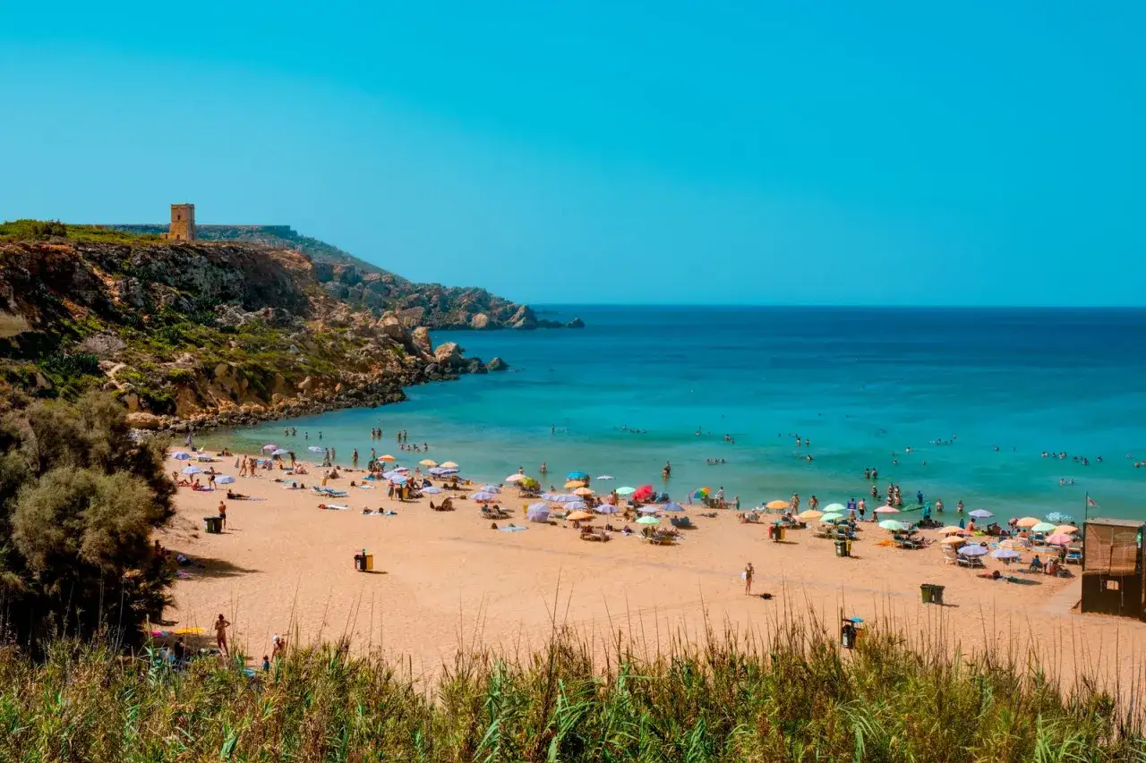Malta plaże piaszczyste z turkusową wodą i ludźmi odpoczywającymi pod parasolami. Na wzg&oacute;rzu widać starą wieżę.