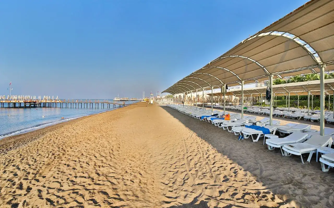 Plaża w Lara, Turcja, z rzędem leżaków pod zadaszeniem i długim molo w oddali.