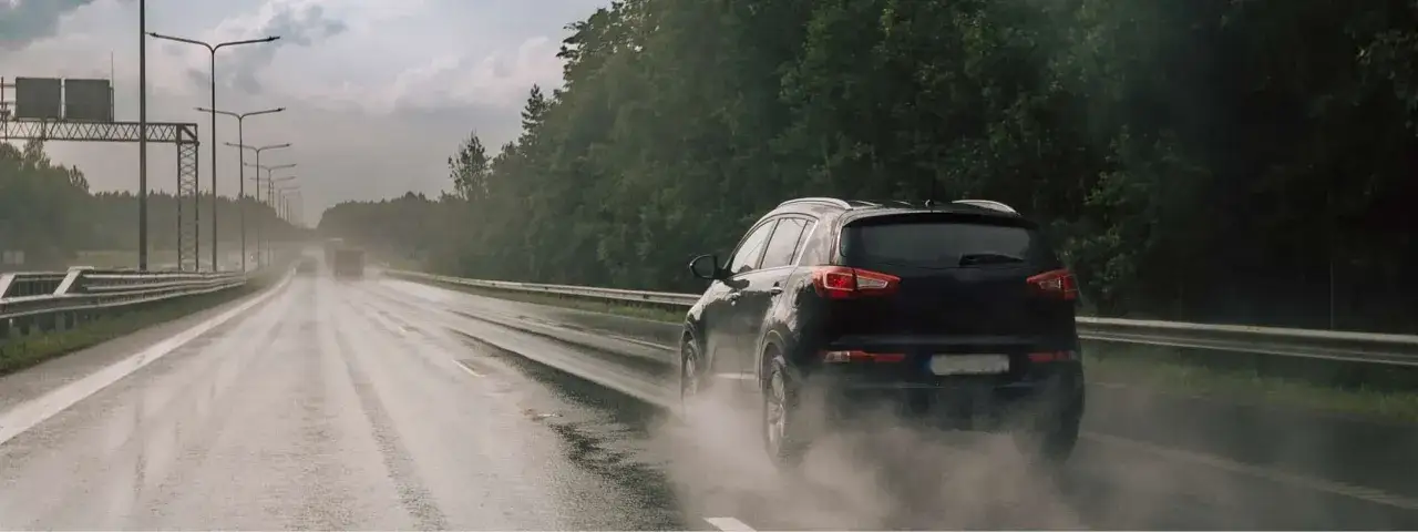 Une voiture noire roule sur une autoroute mouillée, soulevant un nuage de vapeur. La visibilité est réduite, rendant la conduite sous la pluie périlleuse.