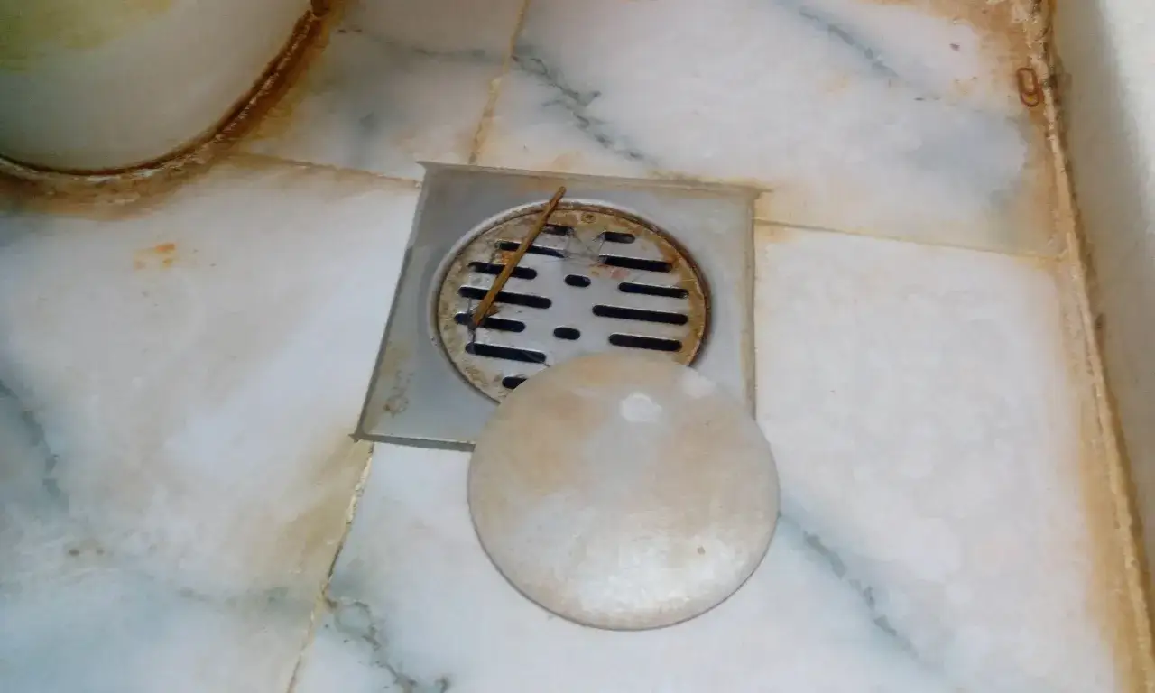 Rejilla de desag&uuml;e con tapa circular y un palillo. El suelo de m&aacute;rmol muestra manchas de humedad y suciedad alrededor del bote sif&oacute;nico.