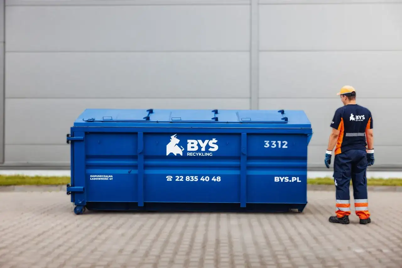 Pracownik firmy BYŚ Recykling stoi obok niebieskiego kontenera. Masz pytanie: gdzie wyrzucić stare płytki ceramiczne? Zadzwoń lub odwiedź BYS.PL.