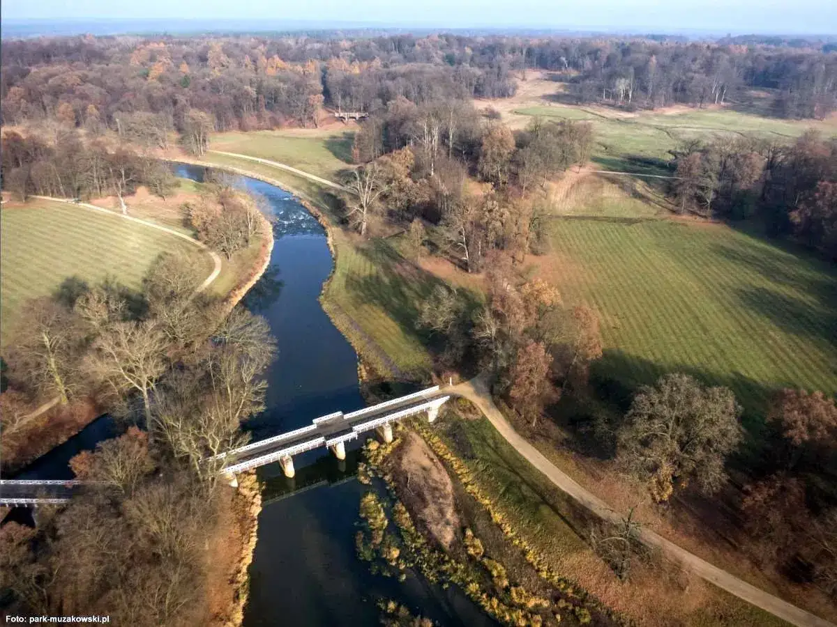 Malowniczy park mużakowski zwiedzanie jesienią. Rzeka przepływa przez park, a biały mostek przecina jej nurt.