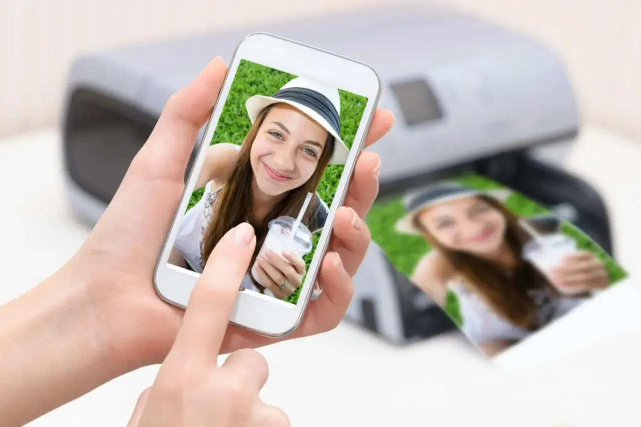 En person håller en mobiltelefon med en bild på en leende tjej med hatt. Bredvid syns en utskrift av samma bild från en skrivare. Enkelt att skriva ut från mobilen!