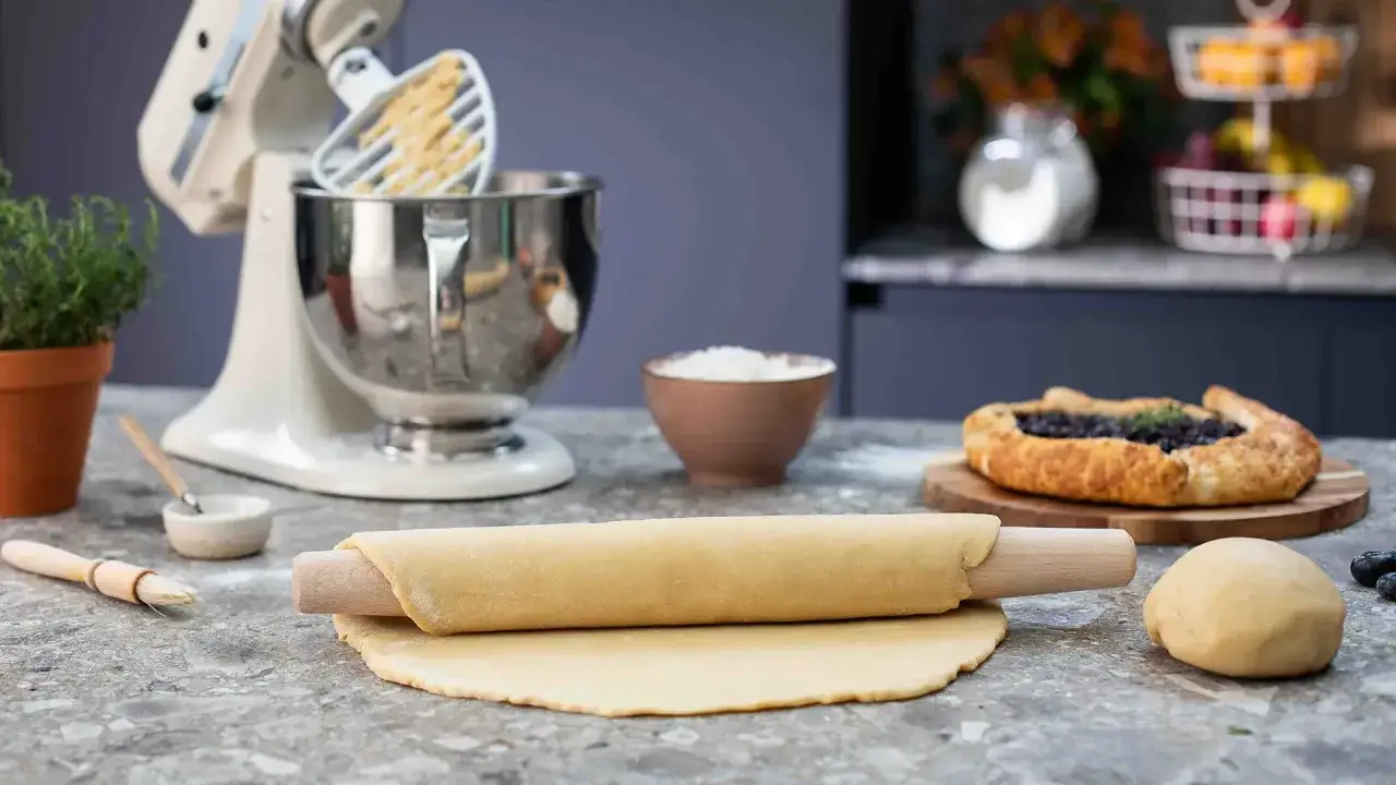 Kavel med pajdeg redo för ett nytt pajdeg recept. En färdig blåbärspaj syns i bakgrunden.