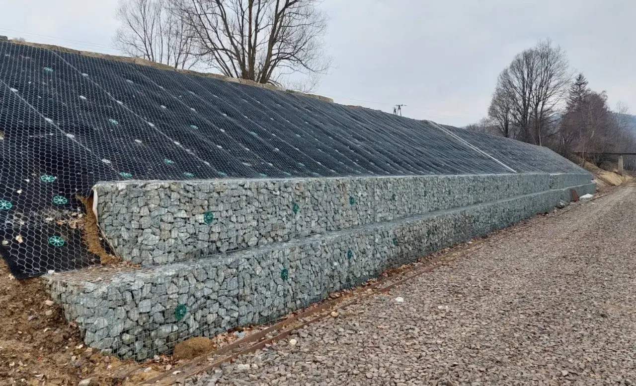 Solidne wzmocnienie skarpy przed osuwaniem: dwie warstwy gabionów z kamienia i siatka geotechniczna na zboczu.