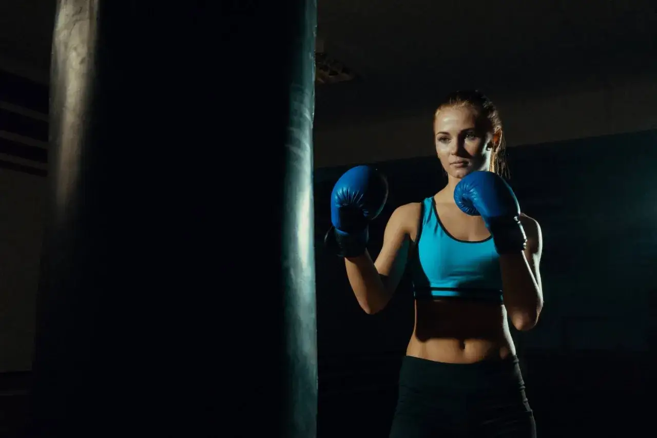 A woman in blue boxing gloves practices her jab punch against a dark punching bag in a dimly lit gym.