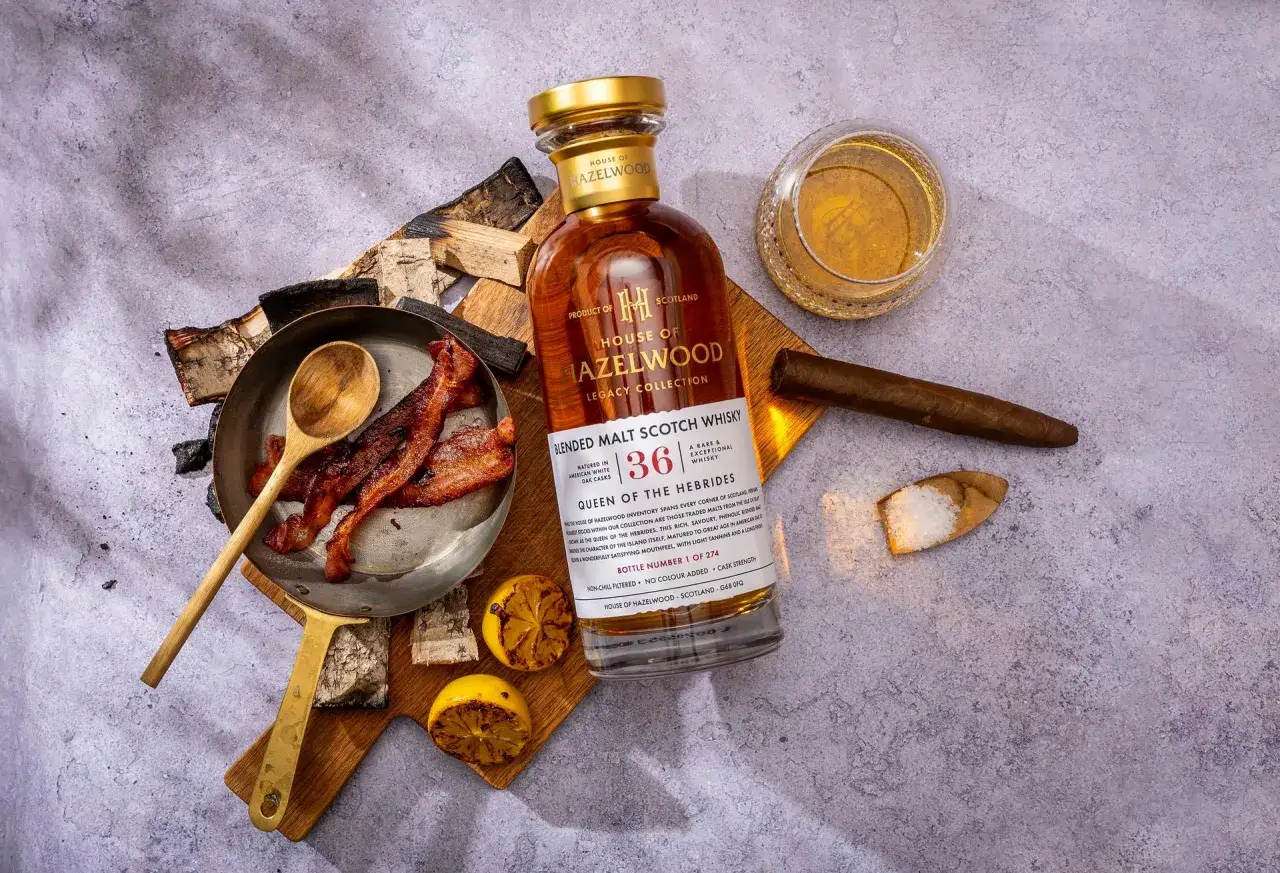 Ein Glas und eine Flasche House of Hazelwood Blended Malt Scotch Whisky, daneben gebratener Speck, gegrillte Zitronen und ein Zigarre.