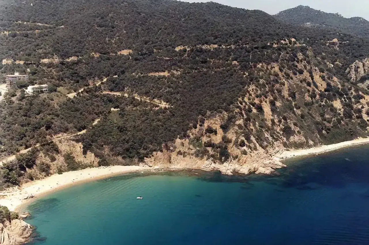 Vista aérea de la platja de Canyerets, con arena dorada, aguas turquesas y una colina boscosa que desciende hasta el mar.