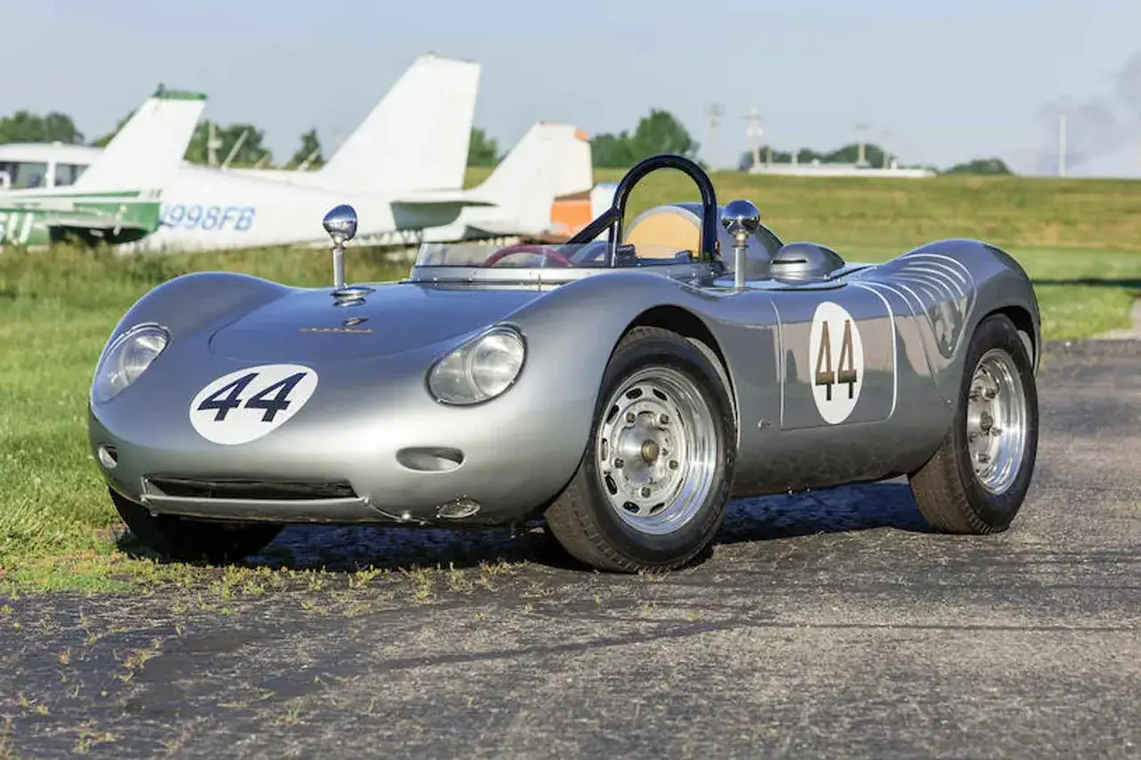 Silberner Porsche 718 RSK mit Startnummer 44, bereit f&uuml;r die Rennstrecke, mit Flugzeugen im Hintergrund.
