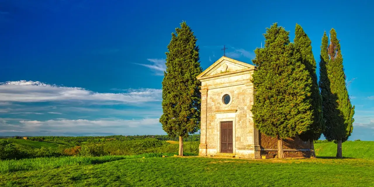 Cappella di San Quirico d'Orcia, circondata da cipressi, su un prato verde sotto un cielo azzurro.