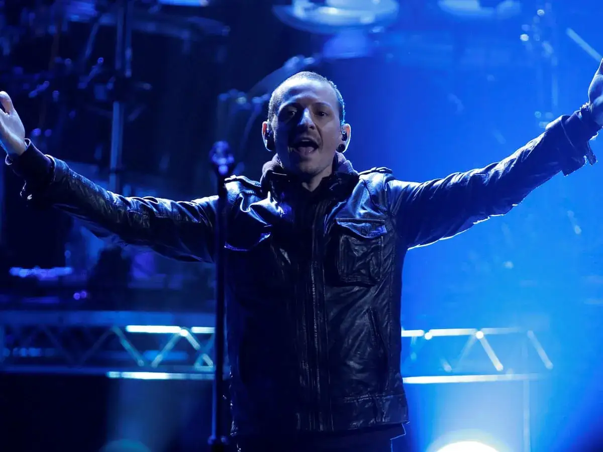 Chester Bennington, wokalista zespołu Linkin Park, śpiewa na koncercie w skórzanej kurtce, z rękami uniesionymi w geście.