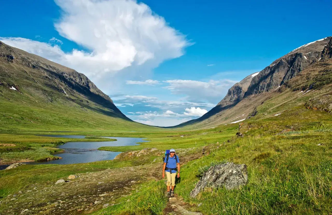 Vandrare på stig i en grön dal med sjö och berg. Syterskalet Hemavan bjuder på storslagen natur.