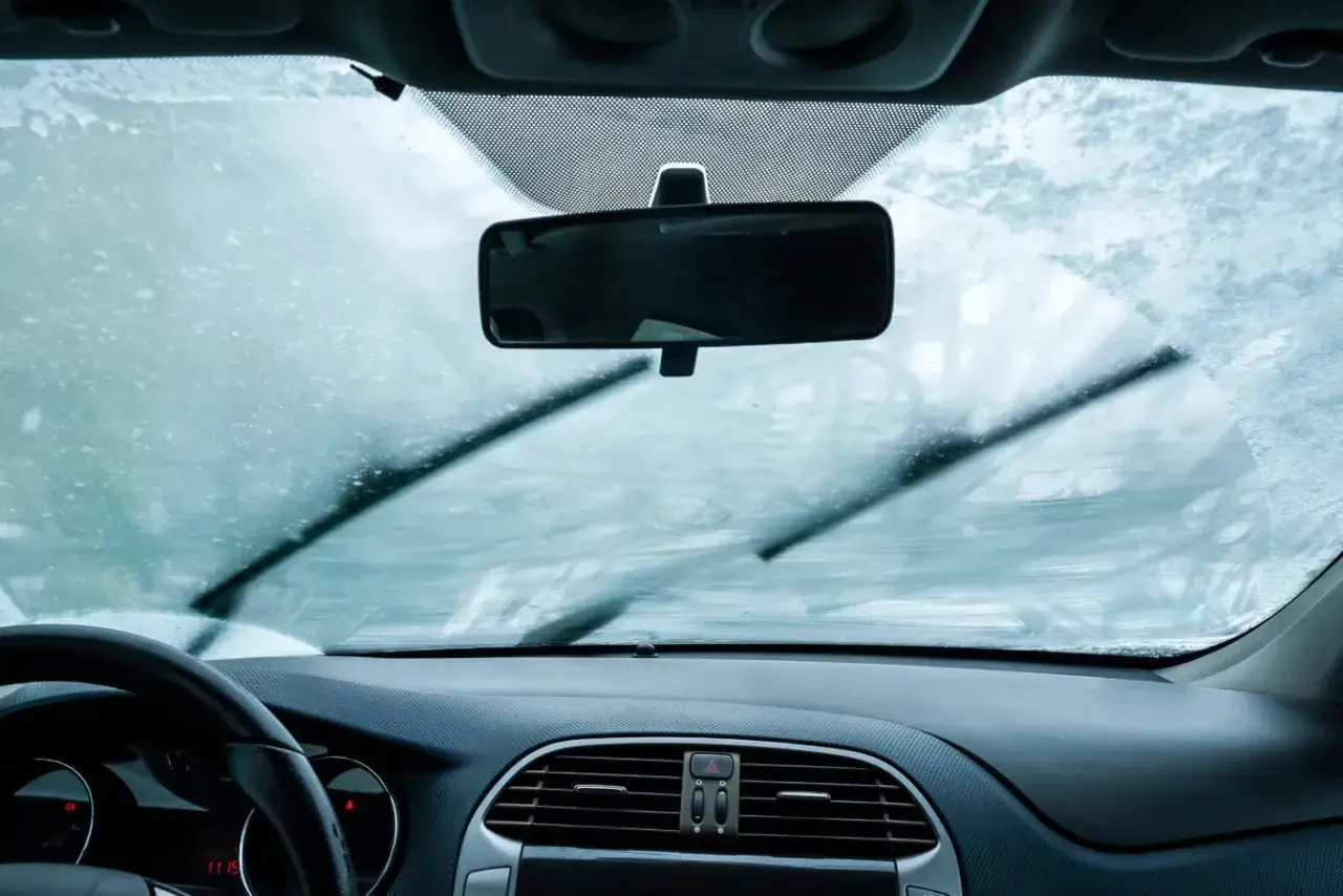 Widok z wnętrza samochodu na zamarzniętą szybę. Wycieraczki próbują oczyścić widok, co przypomina o tym, jak ustawić klimatyzację w samochodzie zimą.