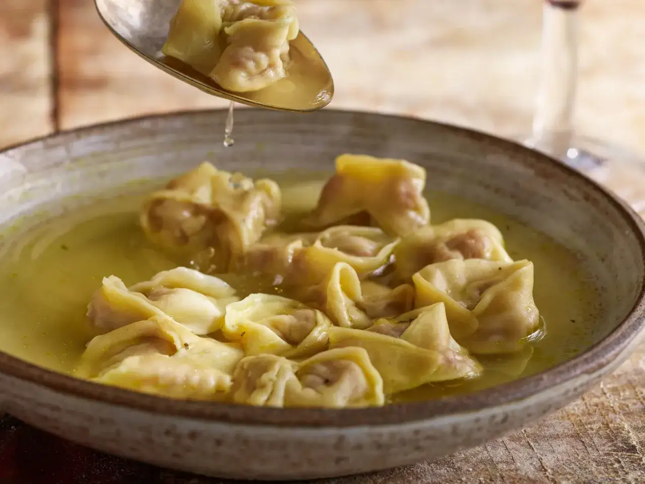 En sked lyfter upp gyllene tortellini in brodo ur en rustik skål.
