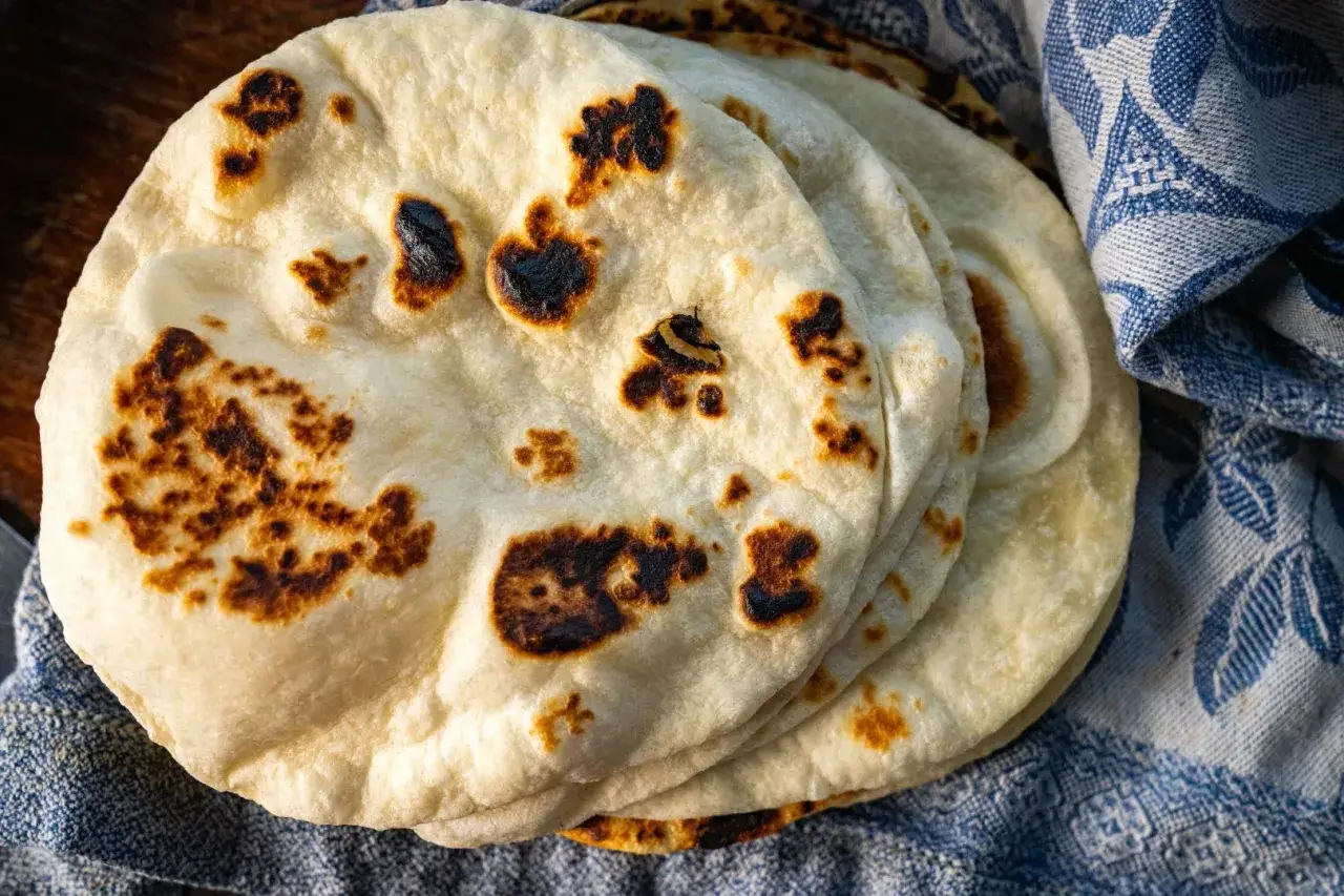 Tortillas de harina recién hechas, doradas y con manchas tostadas, apiladas sobre una tela azul con estampado.