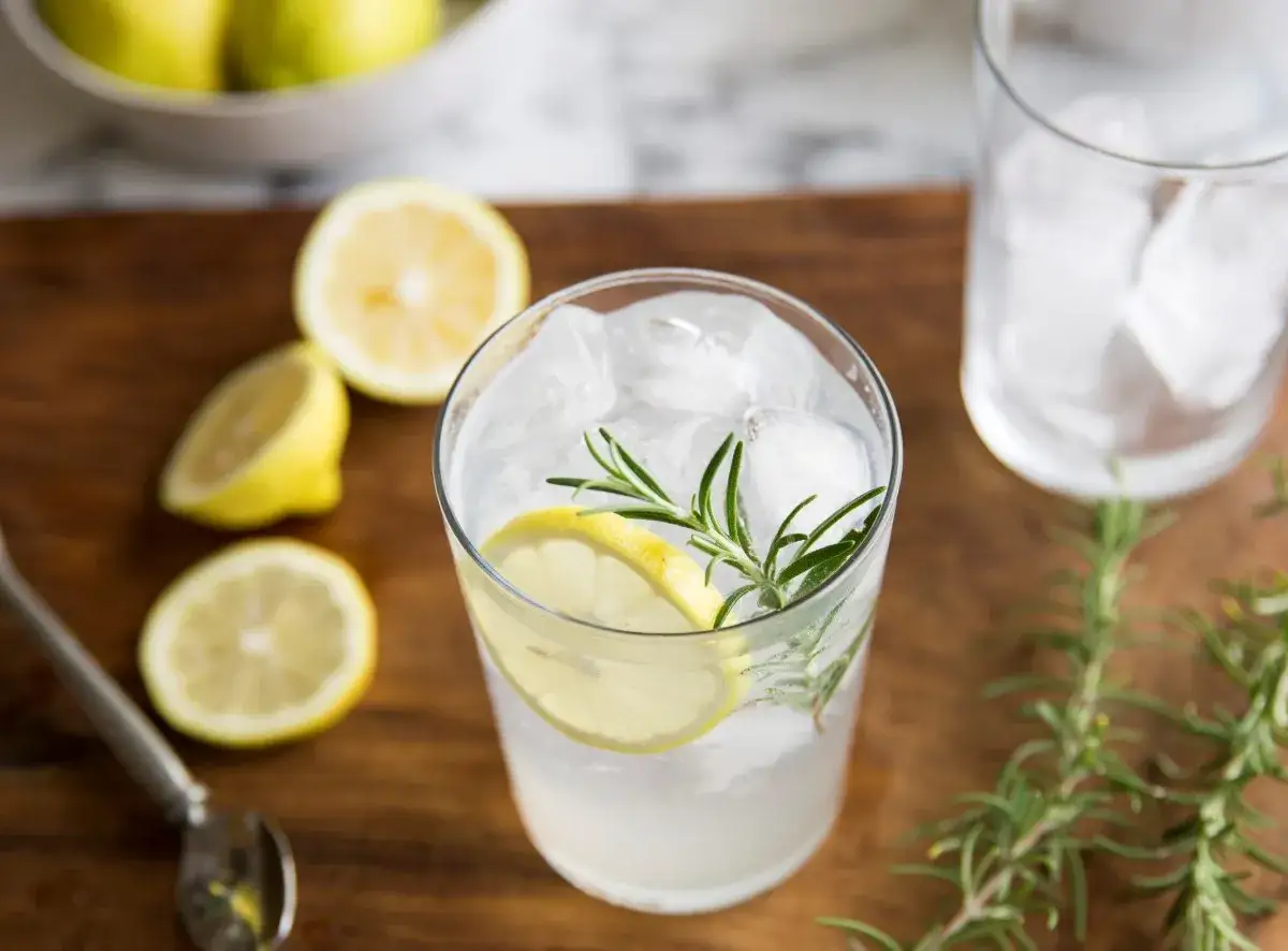 Orzeźwiający gin z tonikiem z plasterkiem cytryny i gałązką rozmarynu, otoczony świeżymi cytrynami i lodem.