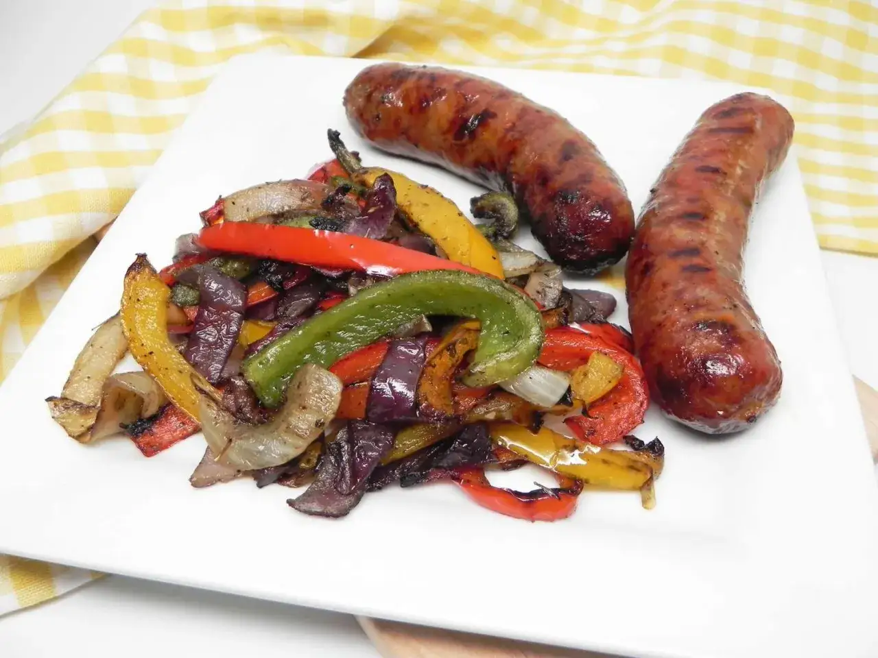Zwei gegrillte Salsiccia liegen neben einem Haufen bunter, gegrillter Paprika und Zwiebeln auf einem weißen Teller.