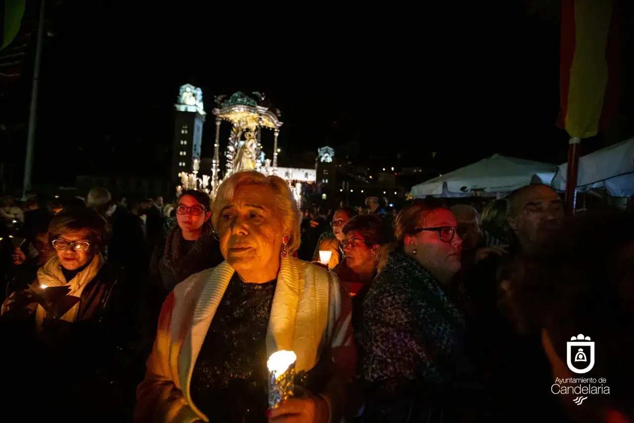 Procesi&oacute;n nocturna con velas. Una mujer mayor, con un chal blanco, sostiene una vela encendida. Al fondo, un paso procesional y gente. Es el santo de candela.