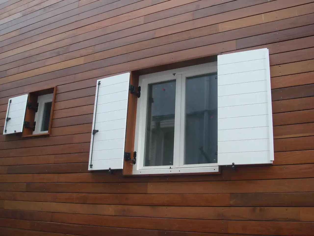 Dos ventanas con contraventanas blancas de listones, una m&aacute;s grande y otra peque&ntilde;a, en una pared de madera oscura.