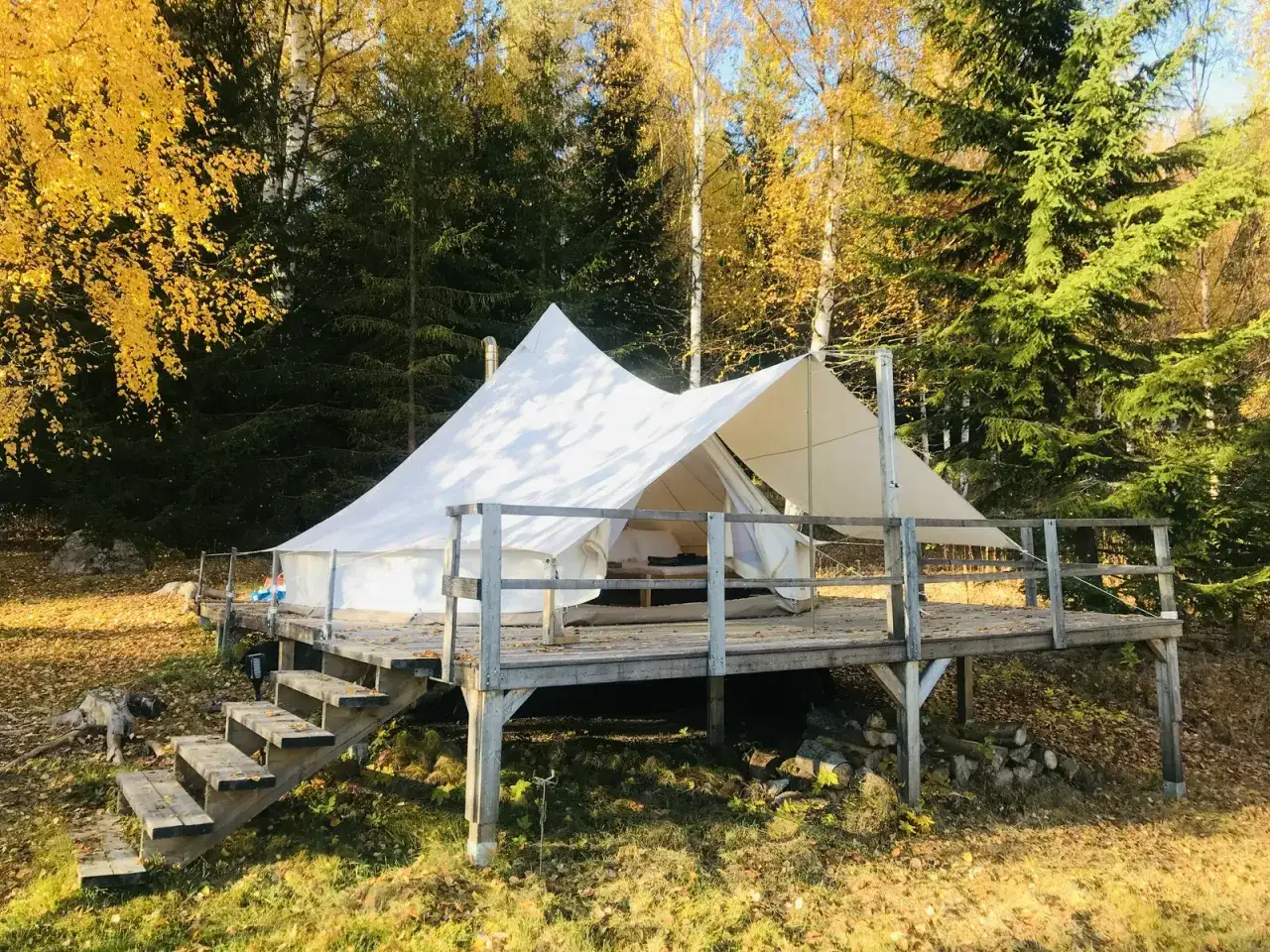 Ett vitt glampingt&auml;lt p&aring; en tr&auml;altan i en h&ouml;stf&auml;rgad skog. Perfekt f&ouml;r att campa i skogen med stil.