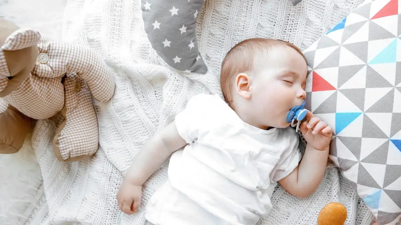 Sover s&ouml;tt med en bl&aring; napp i munnen, omgiven av mjuka leksaker och filtar.