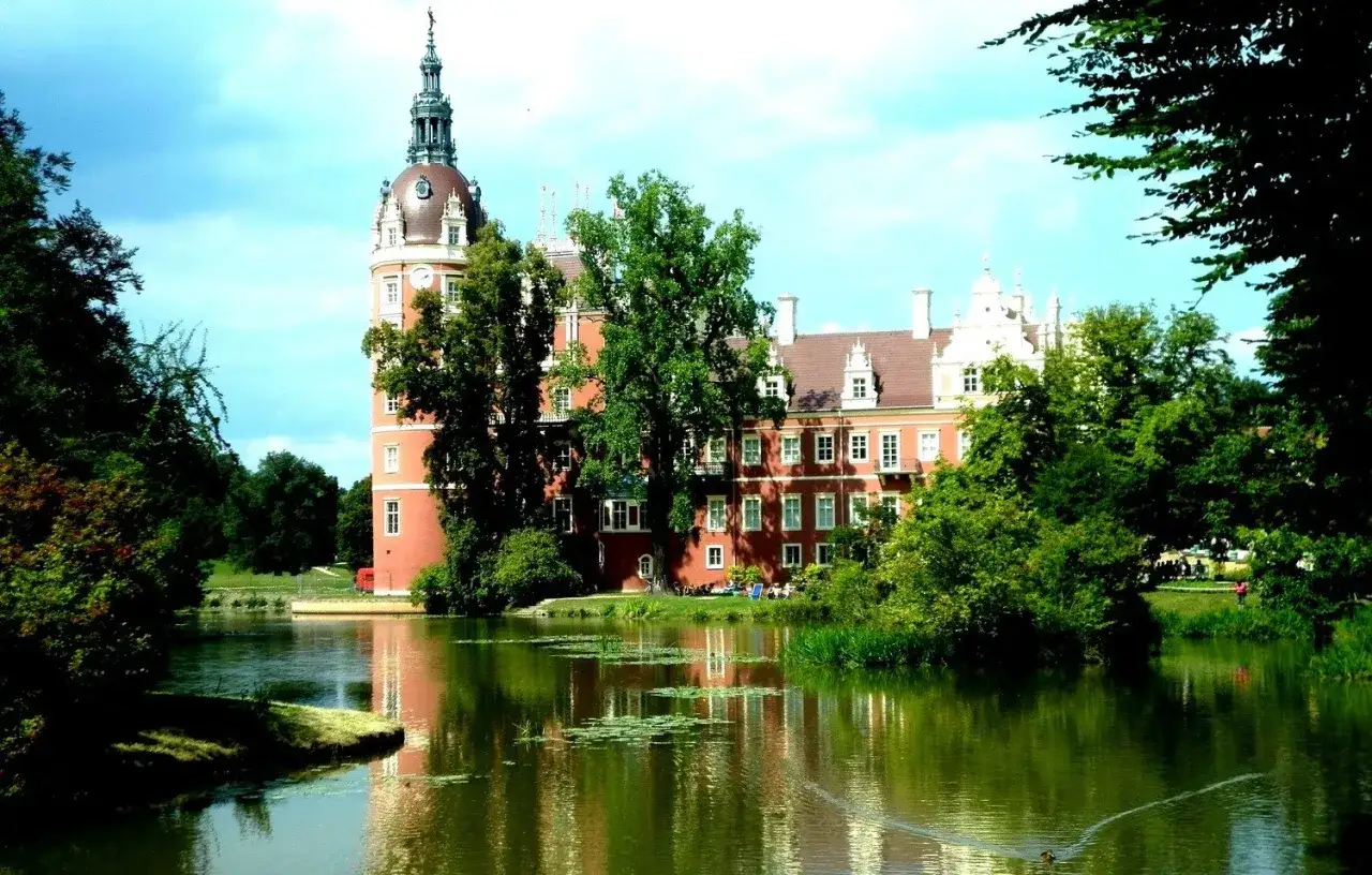 Malowniczy park mużakowski z ceglanym pałacem i wieżą, odbijającym się w spokojnej tafli jeziora.