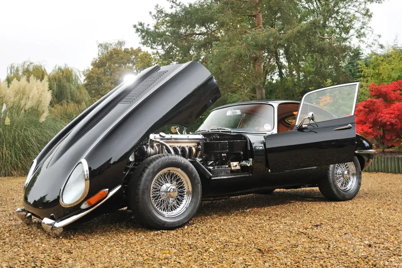 Czarny Jaguar E-Type z otwartą maską i drzwiami, prezentujący potężny silnik. Klasyk motoryzacji na tle jesiennej przyrody.
