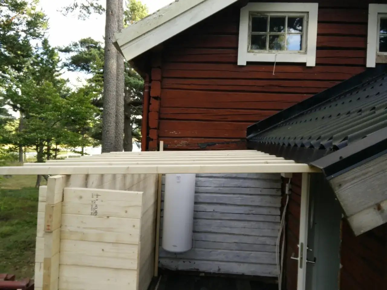 Nybyggd tillbyggnad med takanslutning mot rött trähus. Vattenberedare syns på väggen.