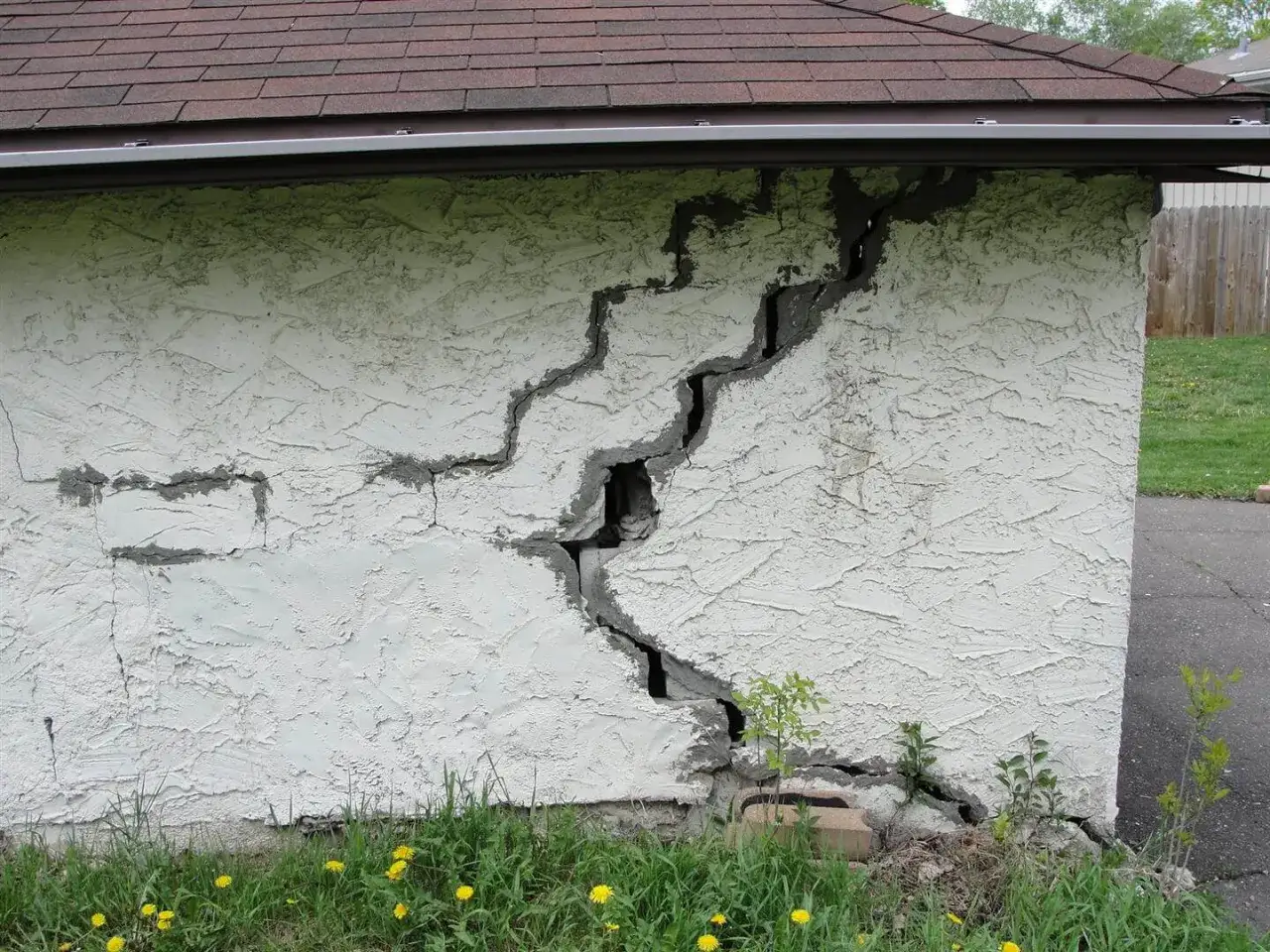A large, jagged crack runs through the stucco wall of a building, revealing the underlying structure. This crack in the concrete slab foundation suggests significant structural stress.