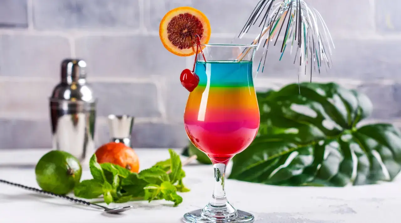 Kolorowe drinki przepisy: tęczowy koktajl z wiśnią i pomarańczą, obok limonka, mięta i shaker.